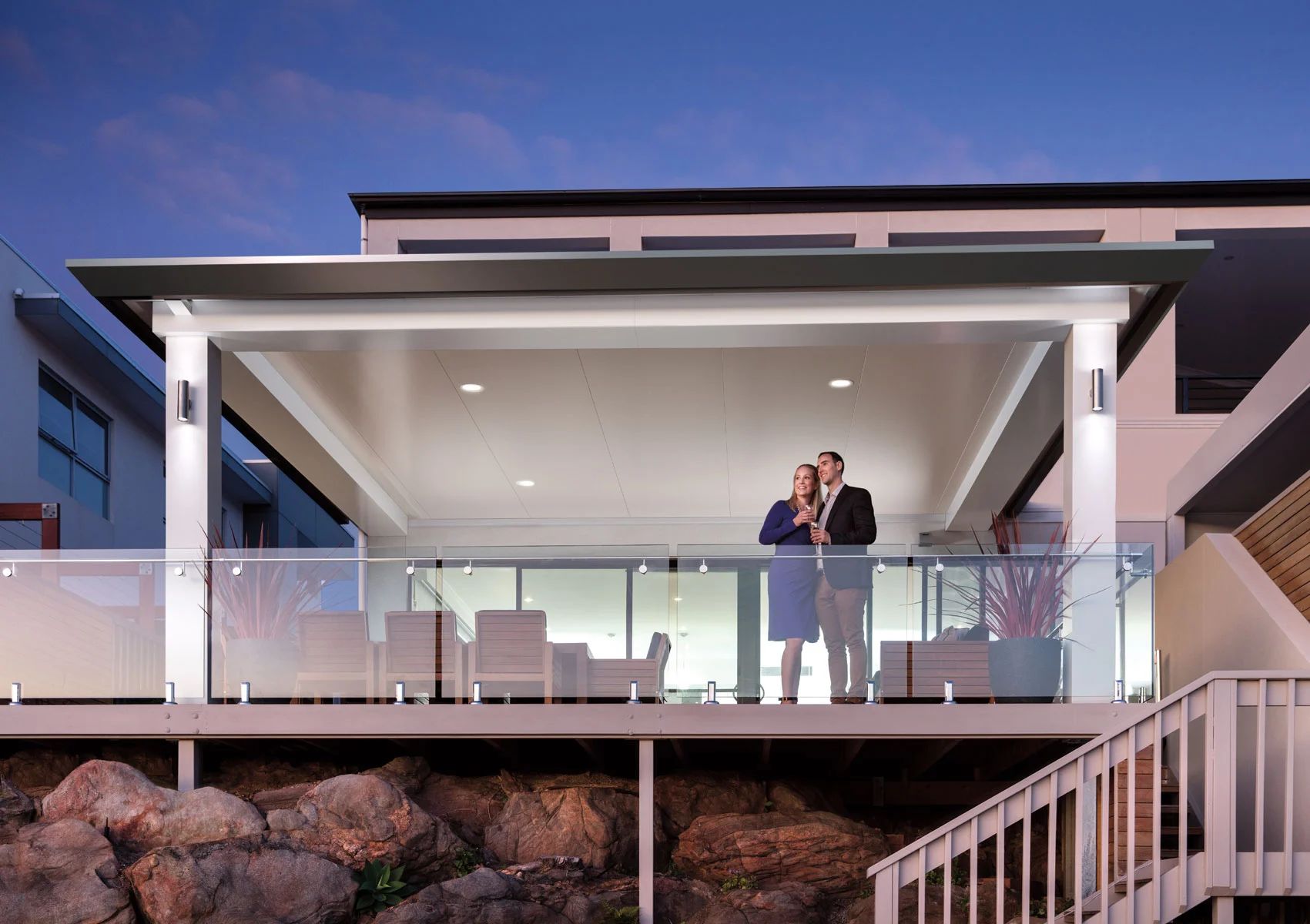 Modern house with glass balcony at dusk, two people standing outside overlooking rocky landscaping