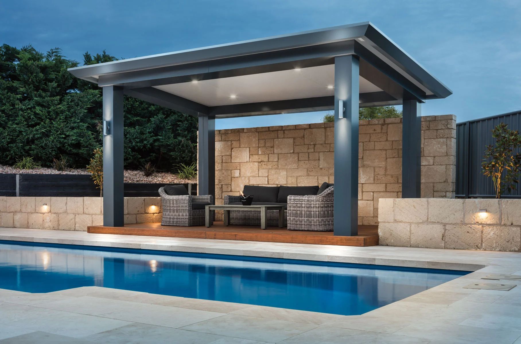Modern backyard poolside cabana with stone wall, lounge chairs, and blue pool at dusk