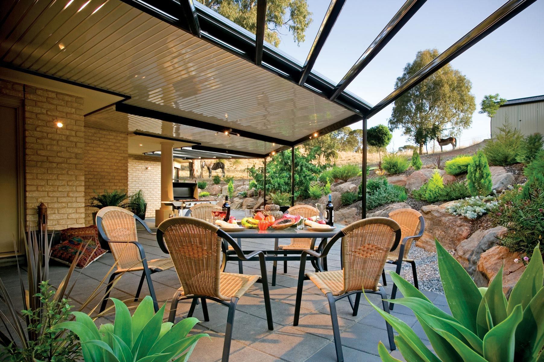 Outdoor patio dining area with wicker chairs, round table, and lush greenery under a slatted pergola.