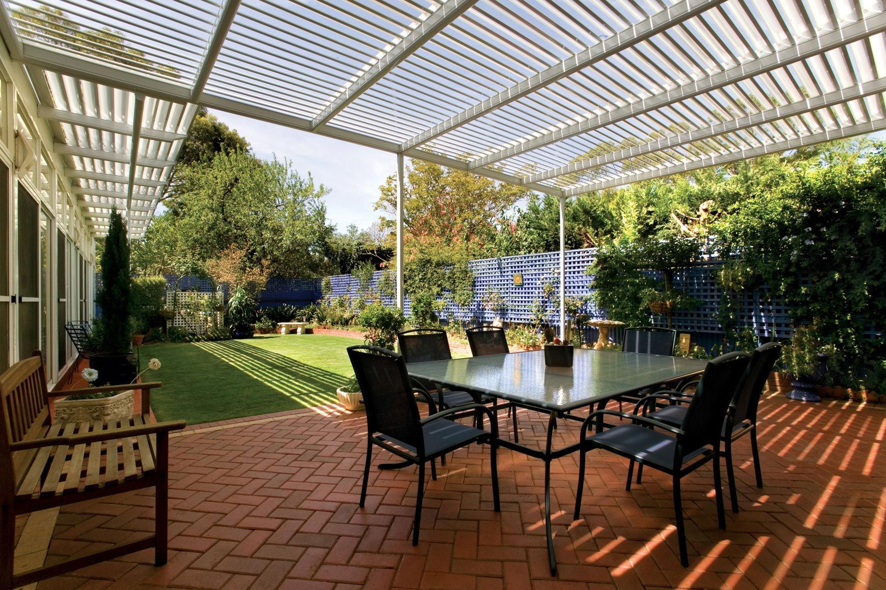 Patio with glass table and black chairs under a white pergola, surrounded by greenery