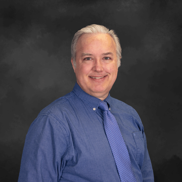 Portrait of a smiling person in a blue shirt and tie against a dark studio backdrop