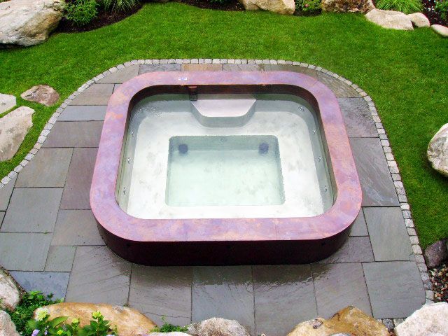 A square hot tub is surrounded by grass and rocks