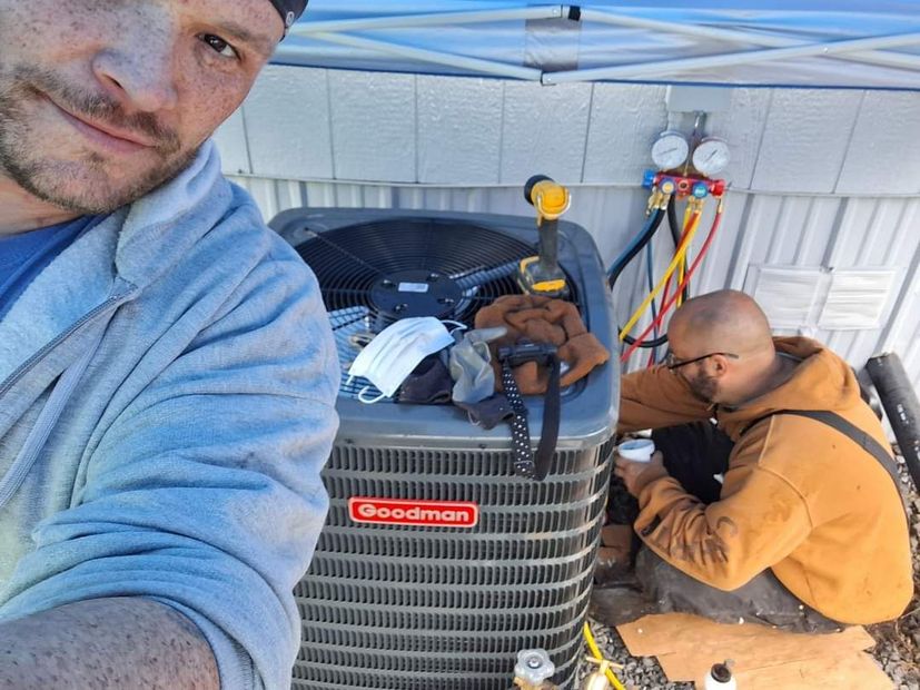 Two men working on an AC unit outdoors. One takes a selfie; the other works on the machine with tools.