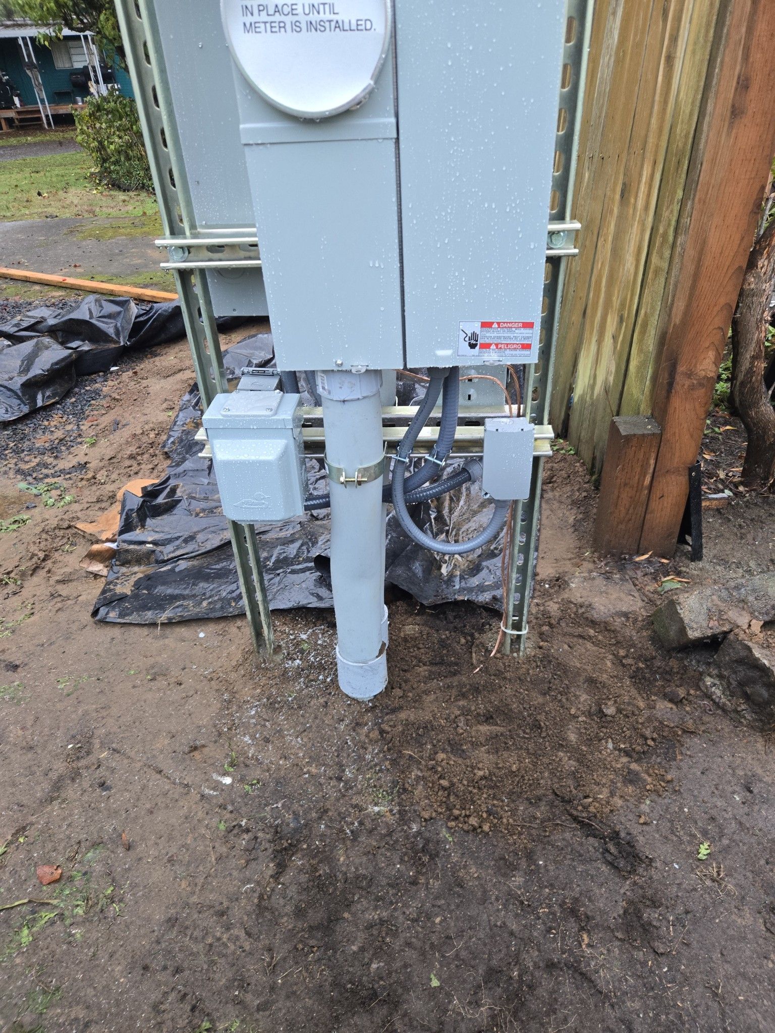 Gray electrical meter box on a pole, mounted on dirt ground next to a wooden fence.