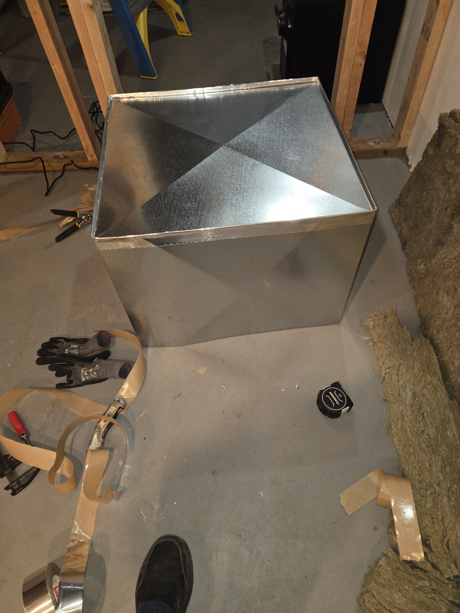 Metal ductwork box on a concrete floor with tools and tape nearby, beside wood framing.