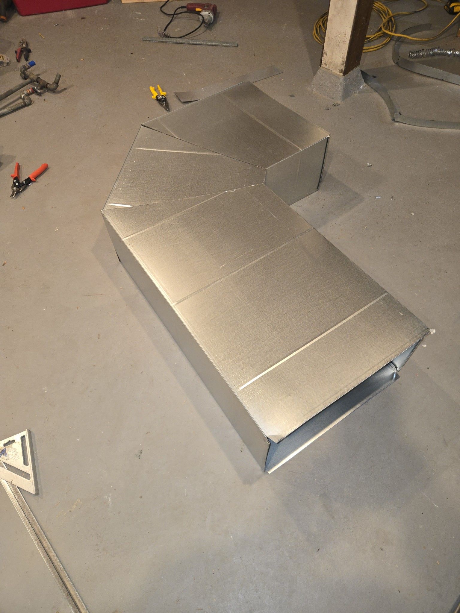 A metal HVAC ductwork elbow lies on a gray concrete floor, with tools scattered nearby.