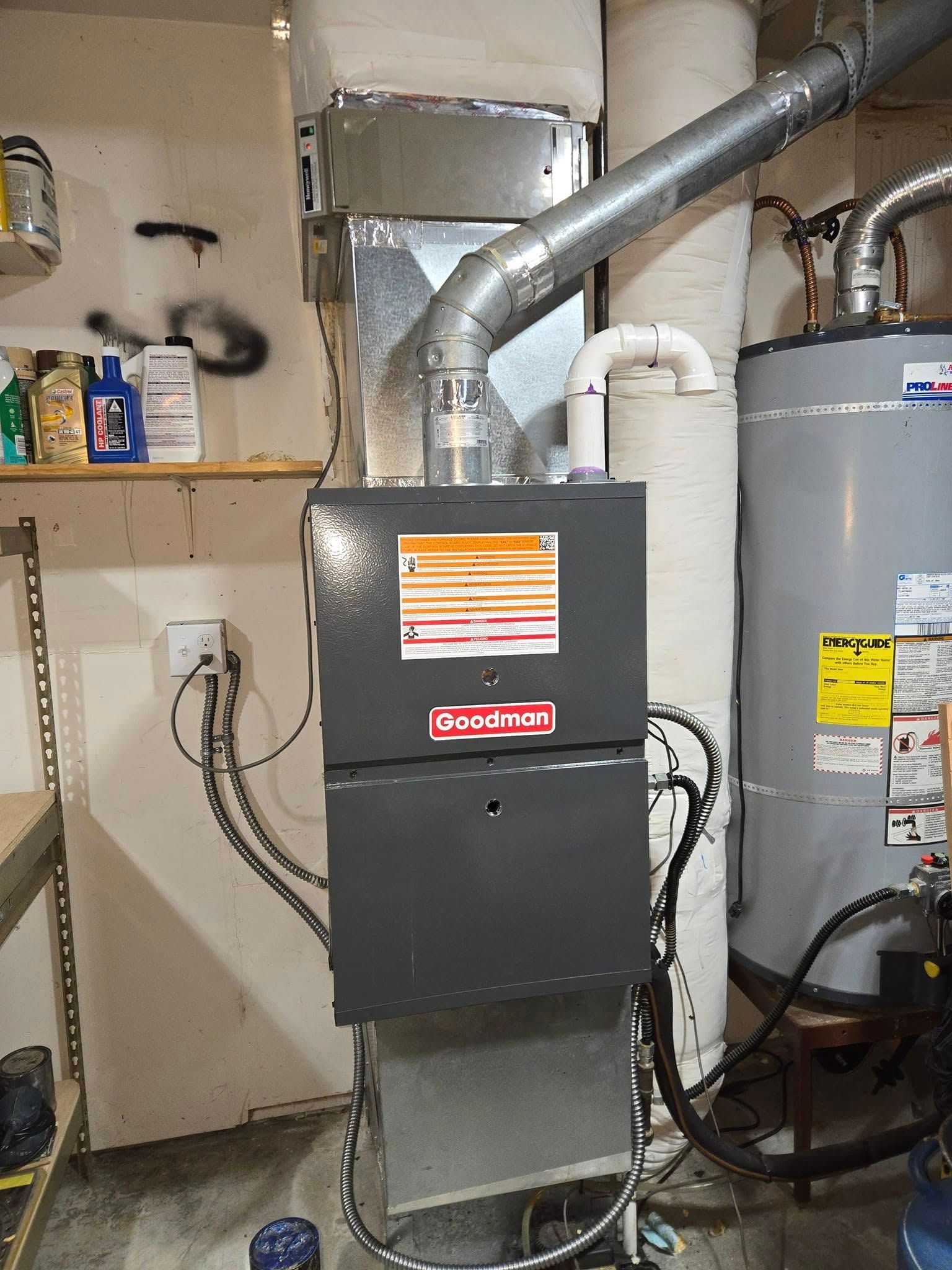 A Goodman furnace and water heater in a utility room. Gray appliances, white pipes, and a small shelf with supplies.