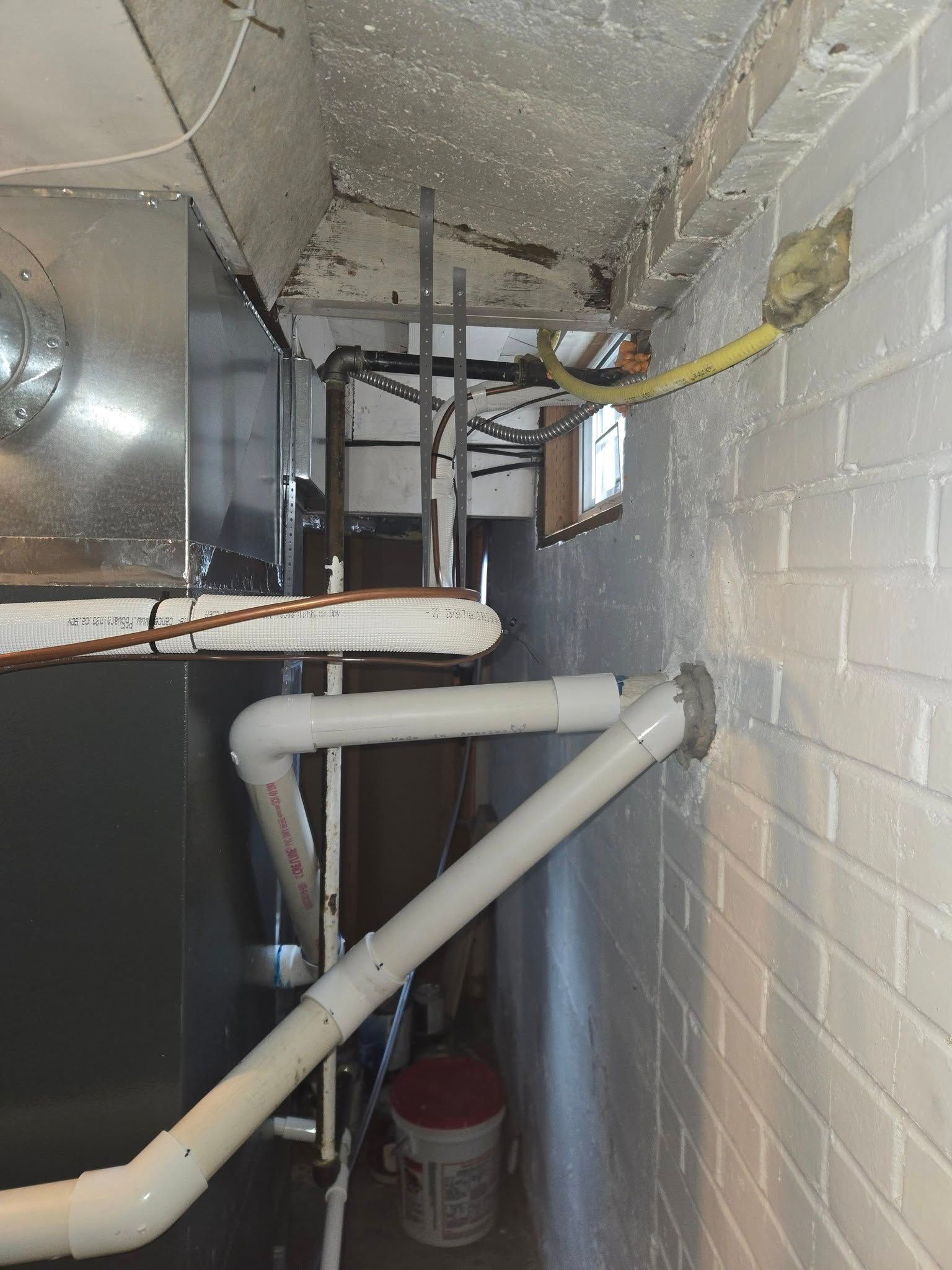White pipes, a furnace, and a yellow gas line against a white brick wall in a basement.