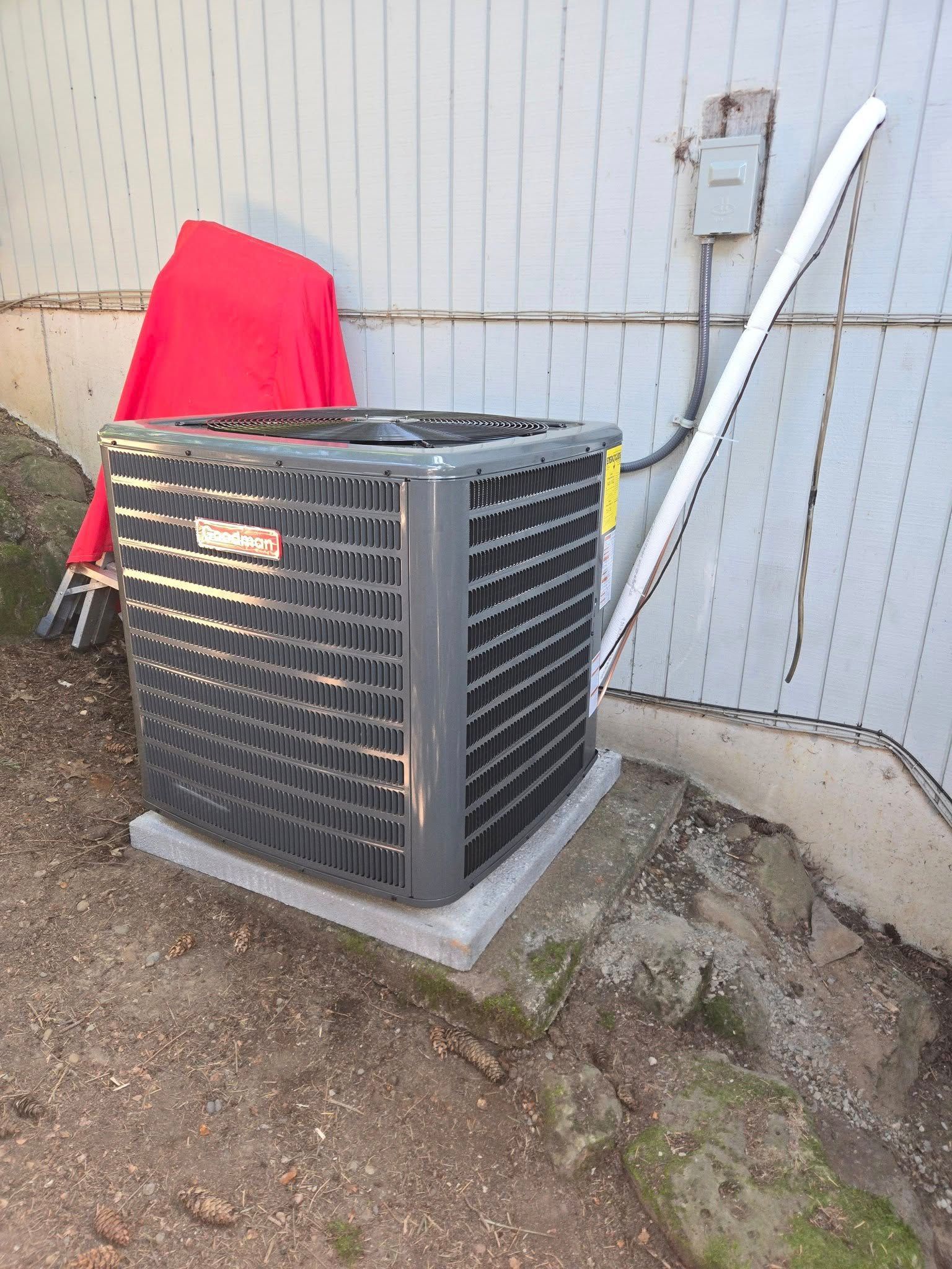 Air conditioning unit on a concrete pad next to a wall, with an electrical box and a red object in the background.