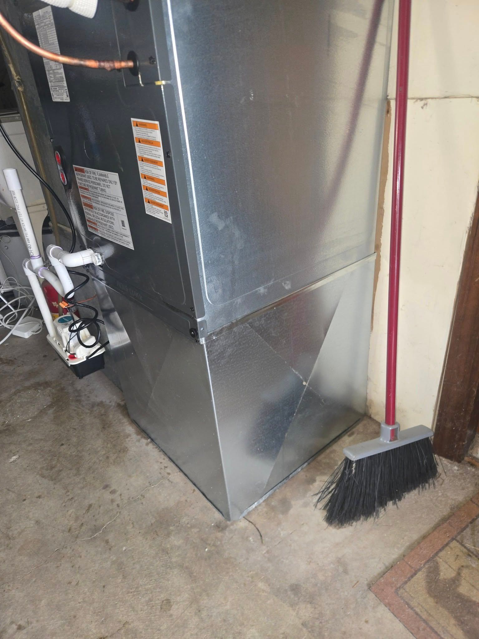 Furnace unit, silver metal, with adjacent broom on concrete floor, indoor setting.