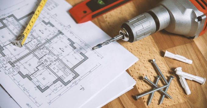 A drill is sitting on top of a house plan on a wooden table.