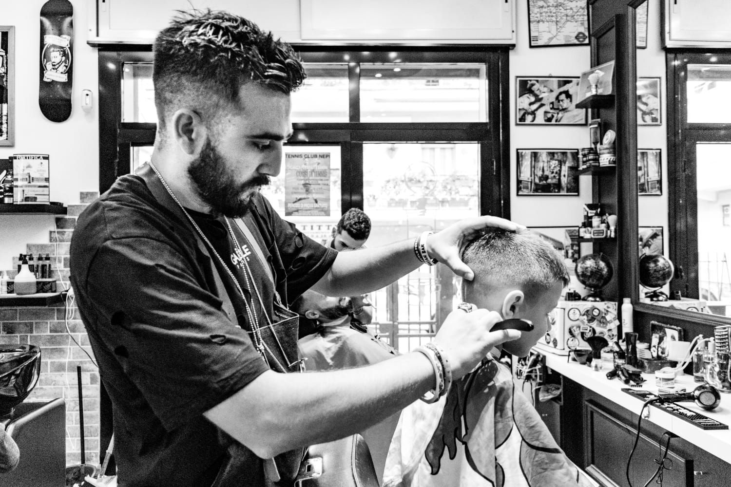 Un barbiere taglia i capelli a un cliente in un negozio affollato, con tosatrici e specchio ben visibili.