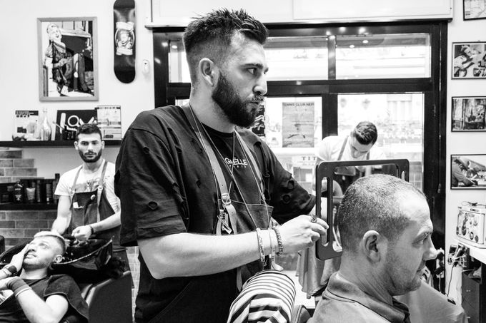 Scena in una barberia: un barbiere taglia i capelli a un cliente mentre altri aspettano sullo sfondo.