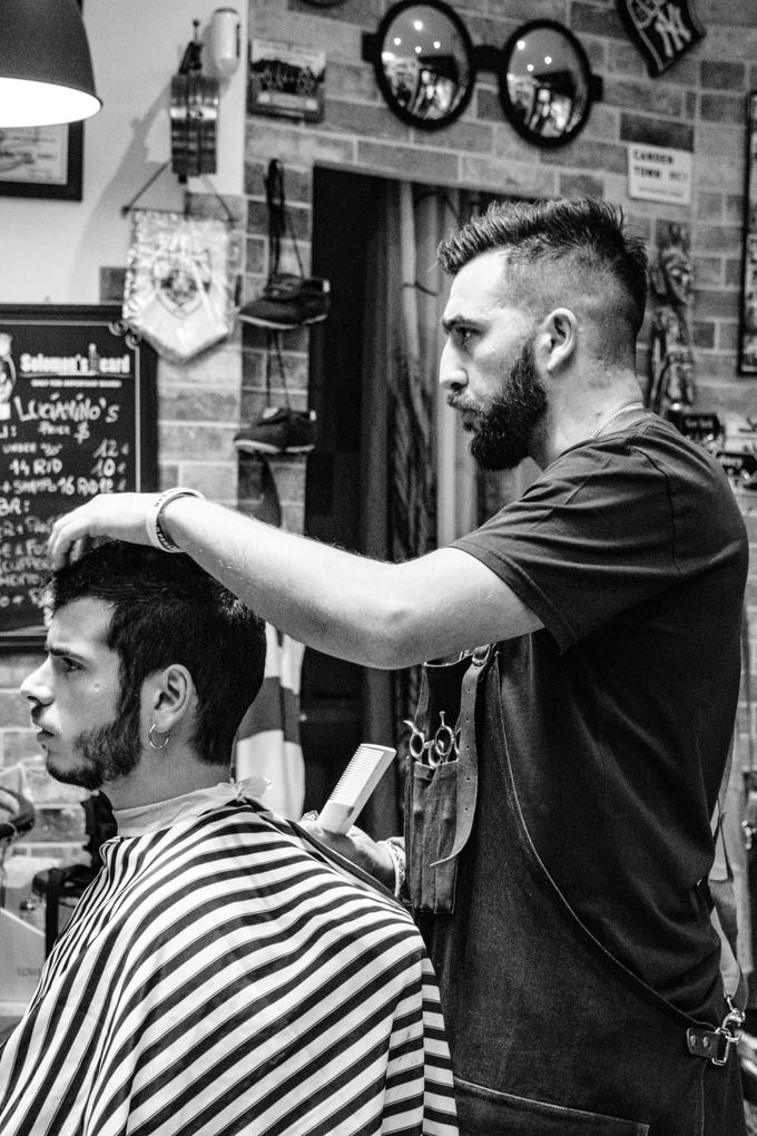 Un barbiere taglia i capelli a un cliente in un negozio, con orologi a muro e decorazioni sullo sfondo.