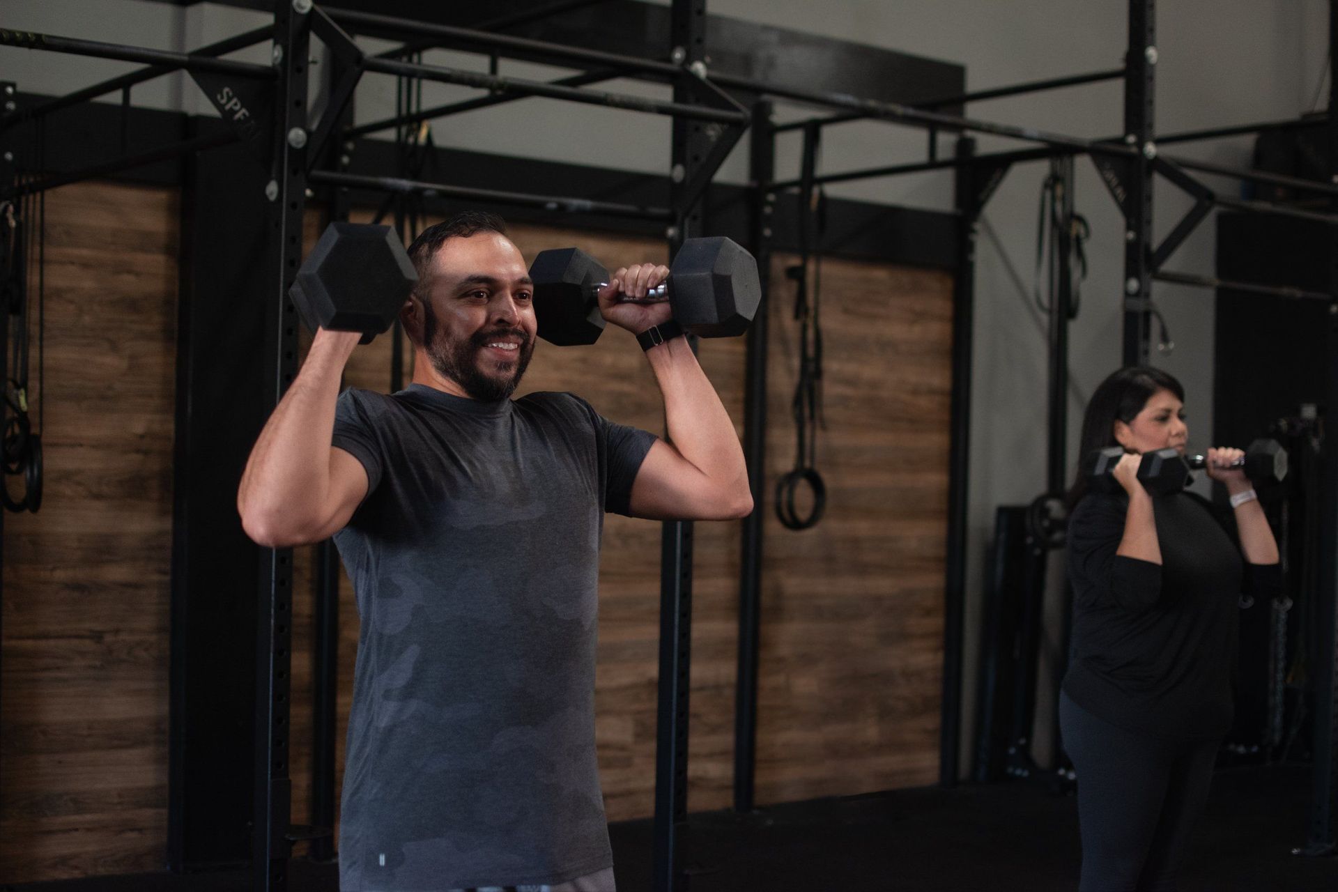 Man and woman lifting dumbbells overhead in a Lubbock gym. Black bars, dark clothes.