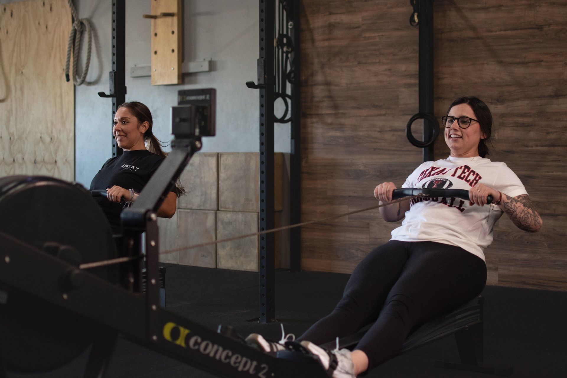 Two people on rowing machines in a Lubbock gym, smiling while exercising. Dark machines, white shirts, dark pants.