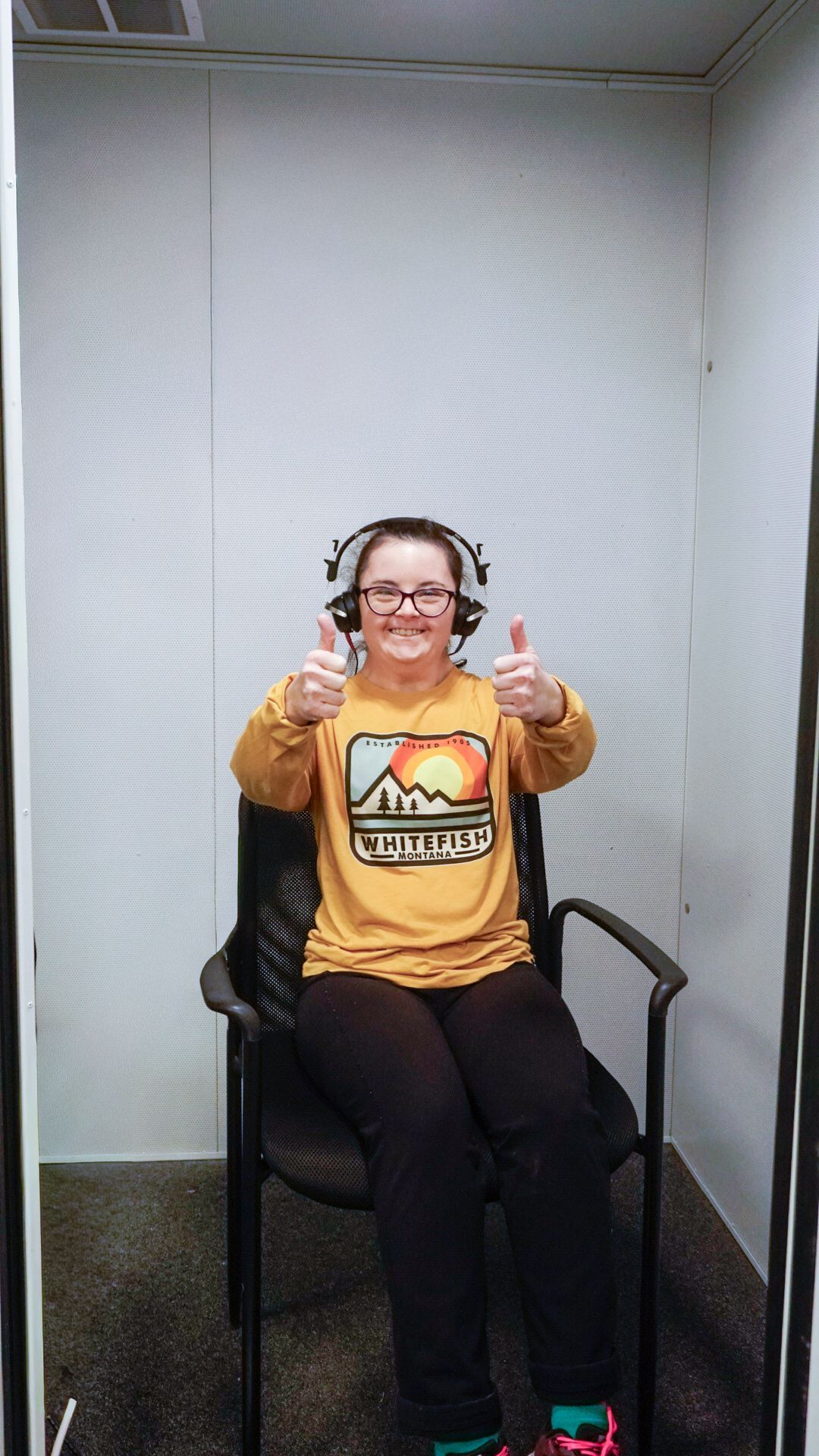 A person wearing headphones and a yellow long-sleeve shirt sits in a soundproof booth, smiling and giving two thumbs up.