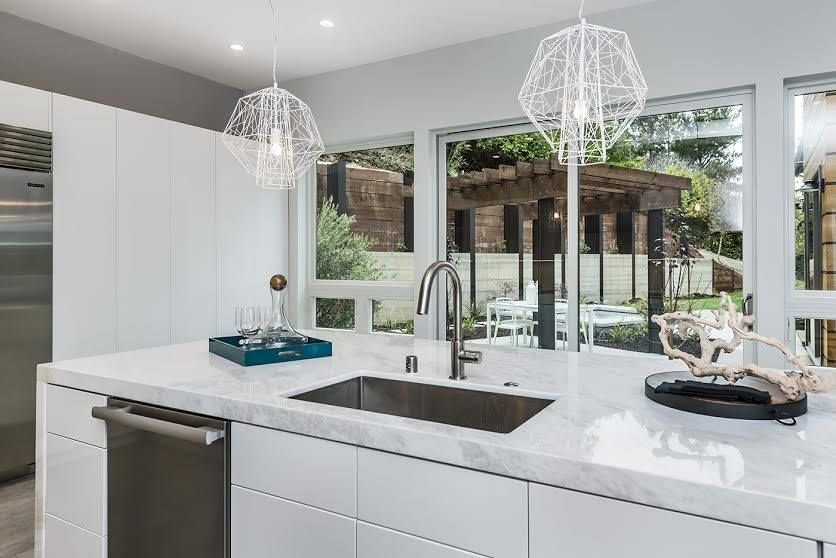 Modern white kitchen with island, pendant lights, and large windows overlooking a patio and garden