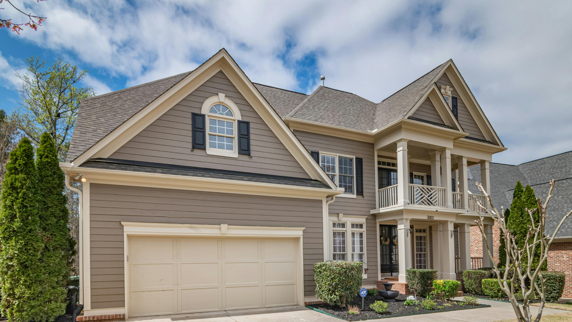 Two-story beige house with a garage, columns, and black shutters under a cloudy sky.