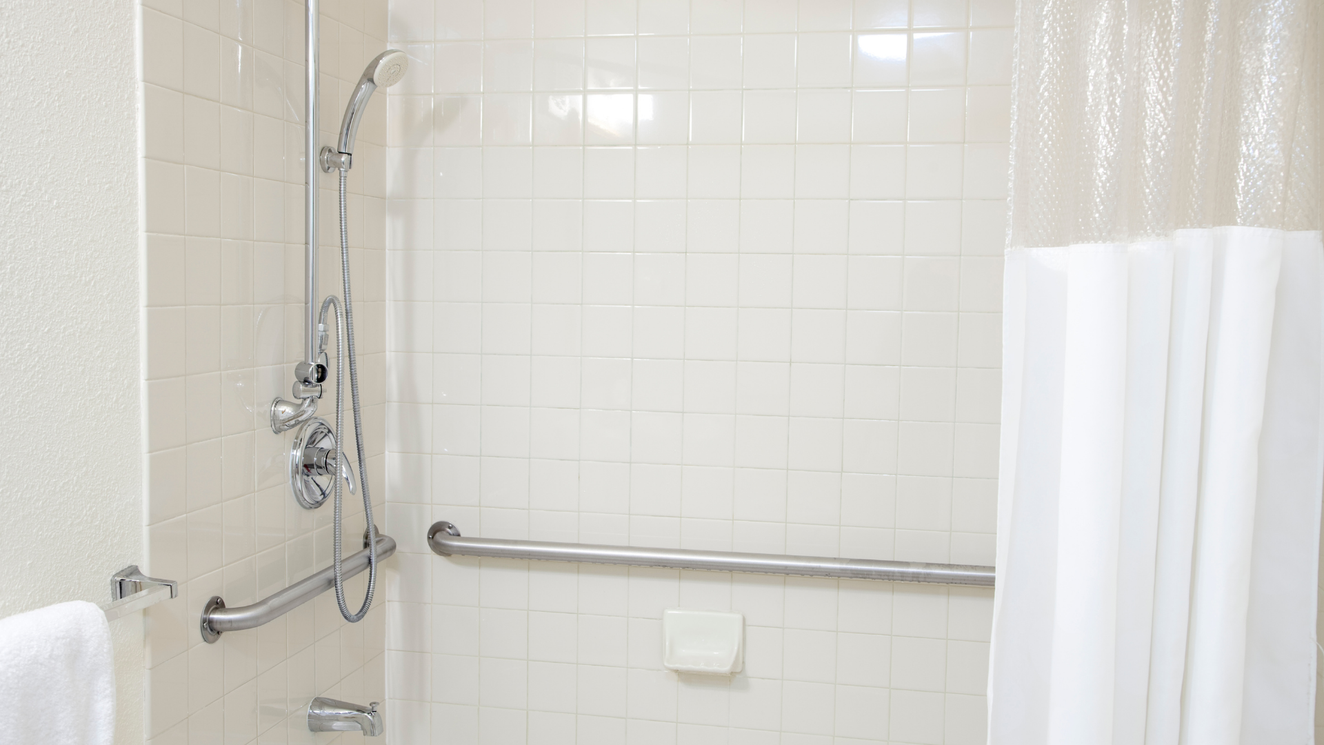White-tiled shower with a handrail, showerhead, and partially open white shower curtain.