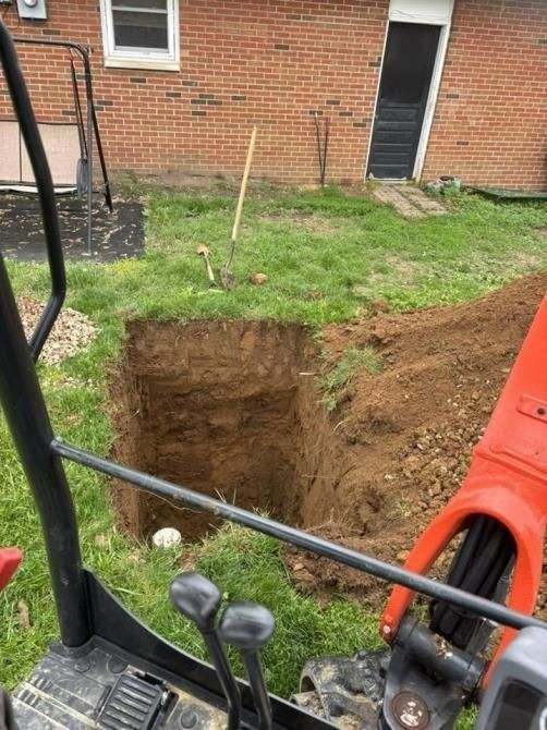 A tractor is digging a hole in the grass in front of a brick house.