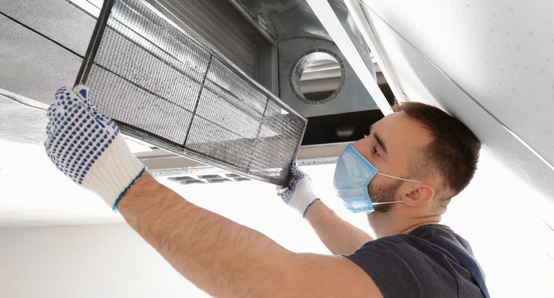 Person with mask and gloves removing a filter from a vent in a ceiling.
