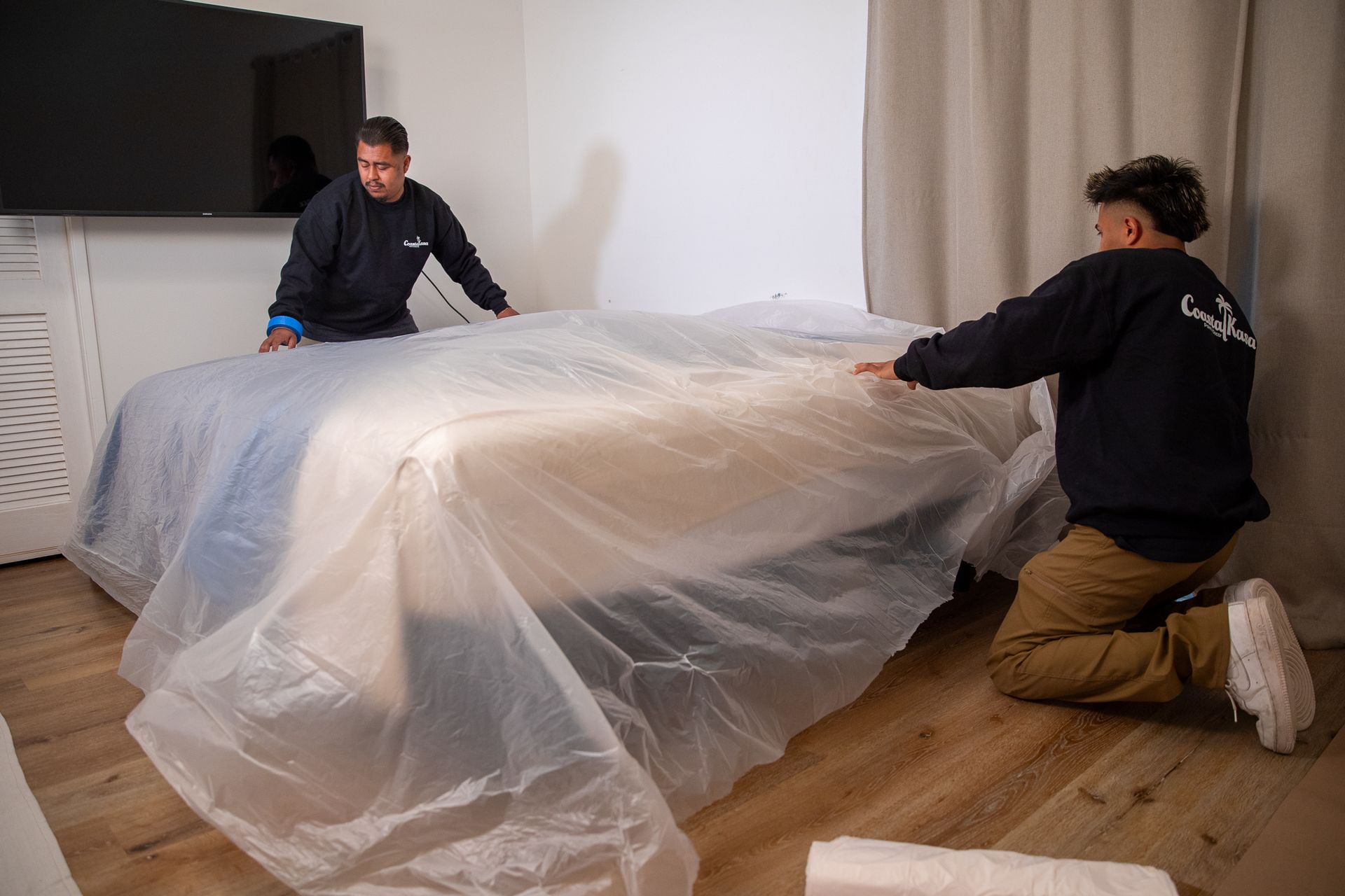 two men are covering a bed with plastic wrap in a living room .