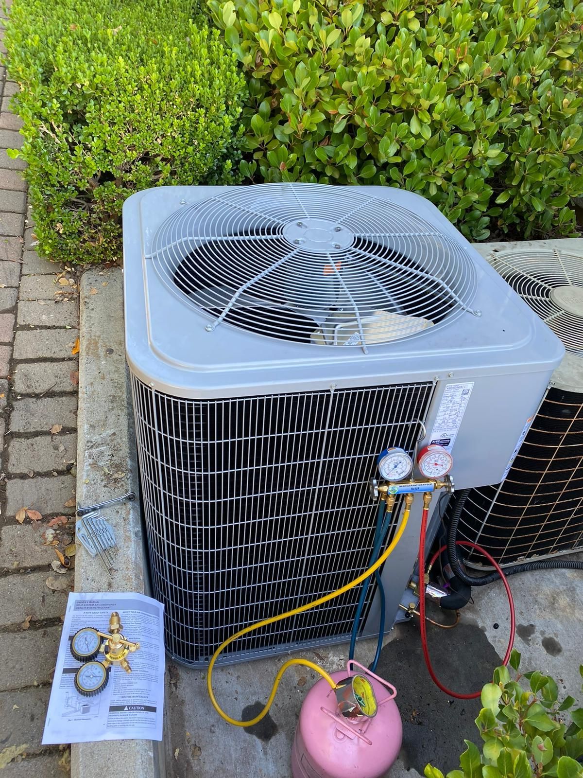 An air conditioner is sitting on the sidewalk next to a bush.