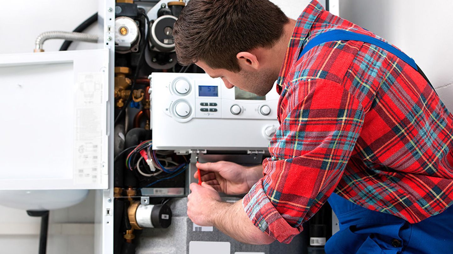 A man in a plaid shirt and blue overalls is working on a boiler.