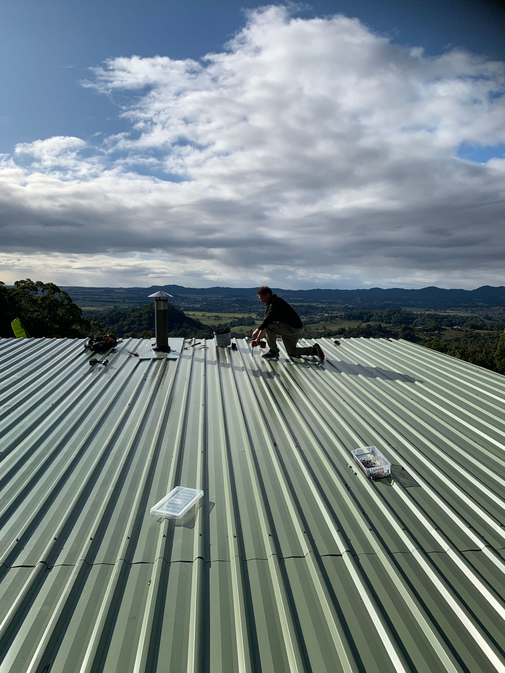 A man is kneeling down on top of a metal green roof — Blue Sky Roofing NSW In Tweed Heads, NSW