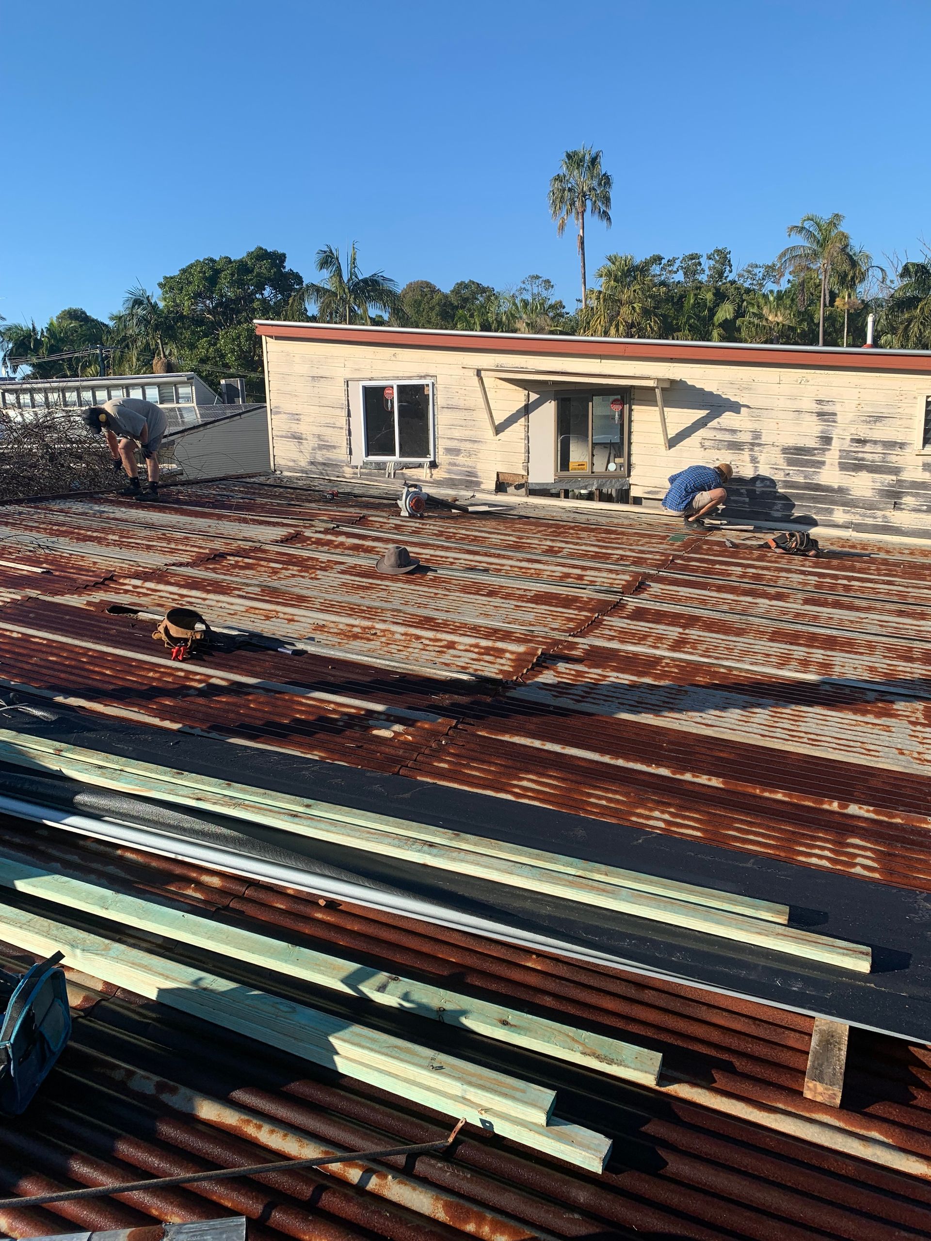 Rusted corrugated iron roof with building in the background, construction materials in the foreground on a sunny day.