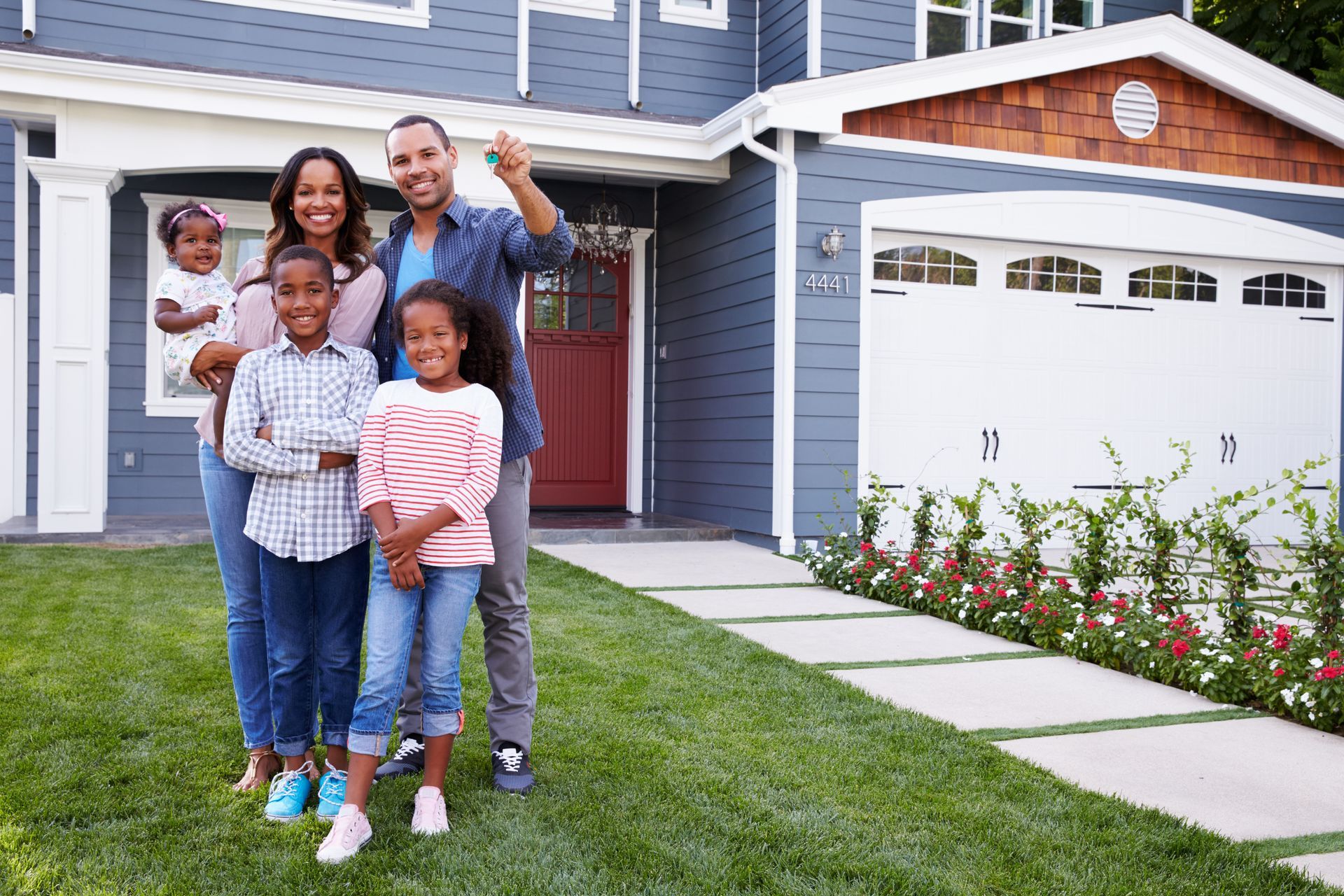 Family of five smiling in front of their new home, holding keys. Blue house, green lawn, sunny day.