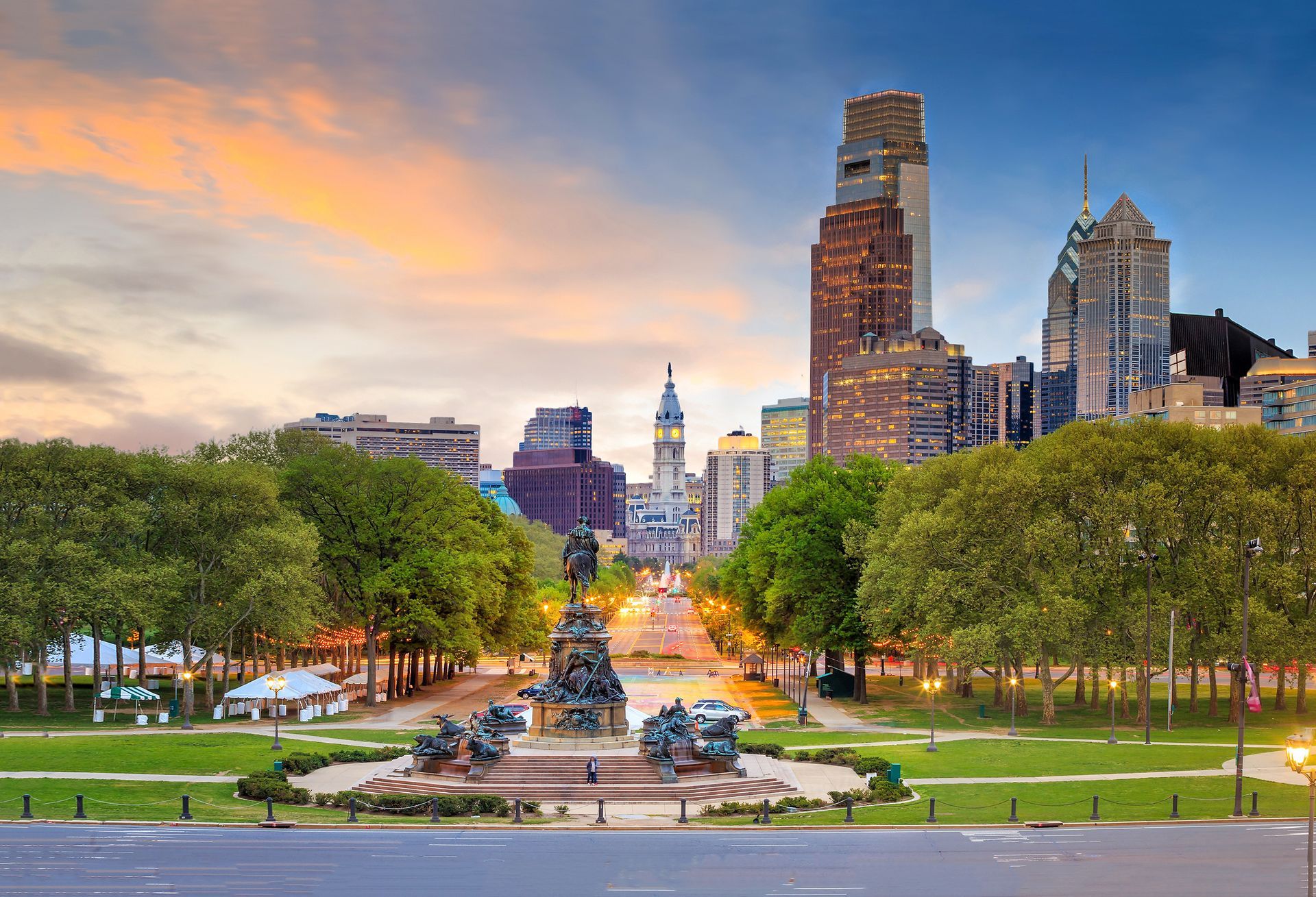 Philadelphia skyline at sunset with a park in the foreground. Buildings, trees, and bronze sculptures are visible.