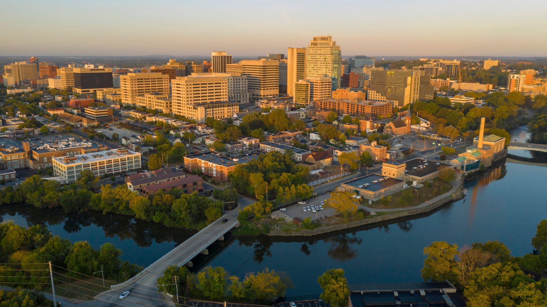 Aerial view of a cityscape at sunset, buildings reflected in river. Golden light bathes the scene.
