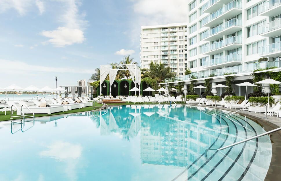 A large swimming pool surrounded by chairs and umbrellas in front of a building.