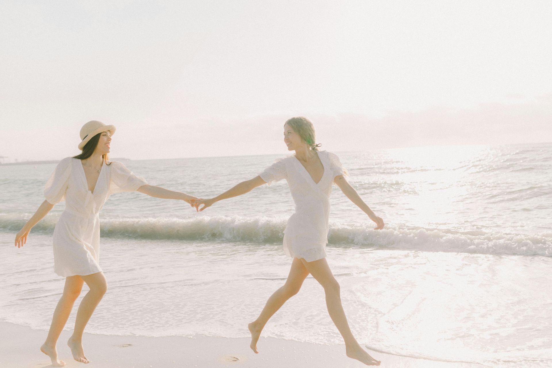Two women in white dresses are holding hands on the beach.