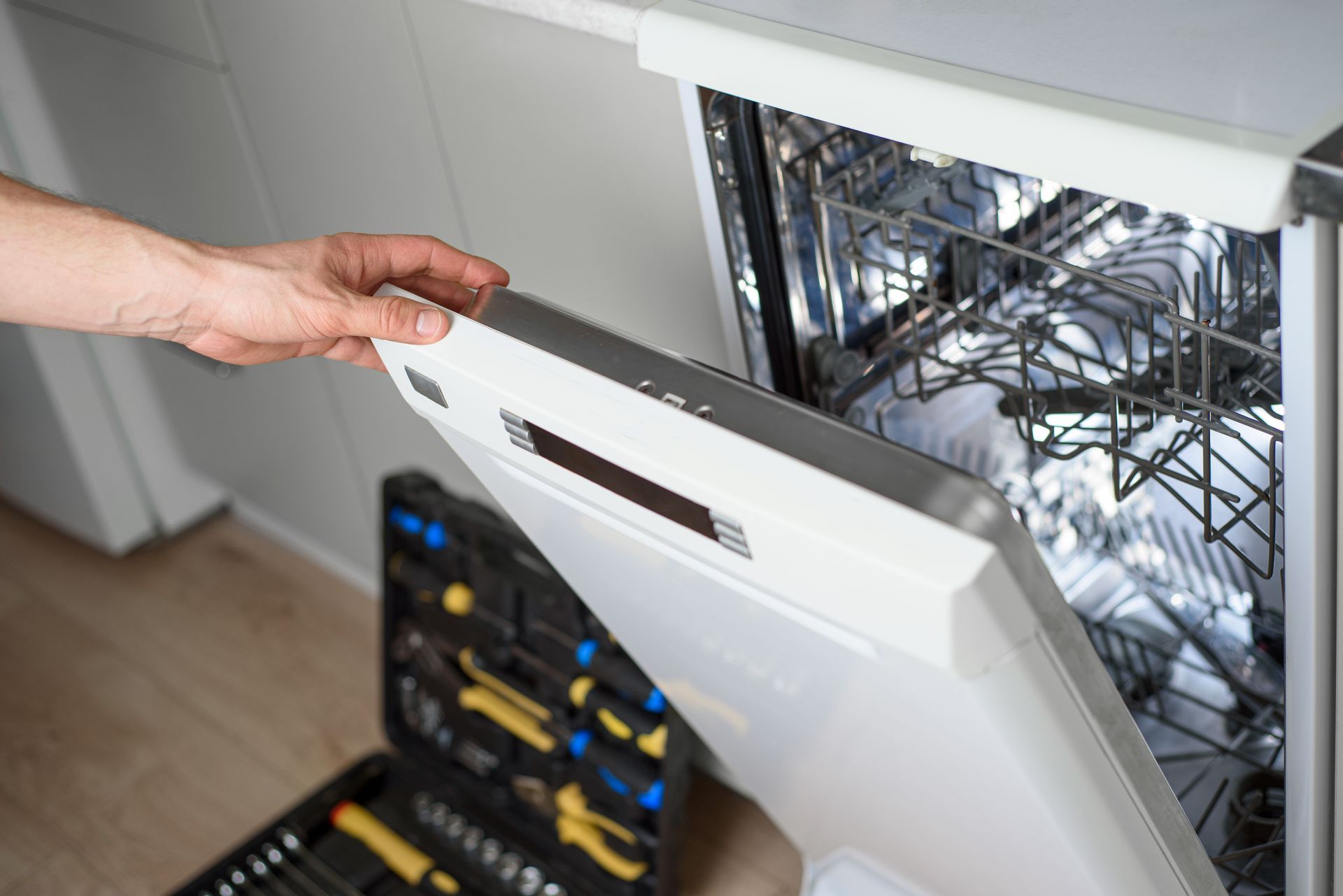 Hand opening a dishwasher door in a kitchen, revealing the wire racks inside