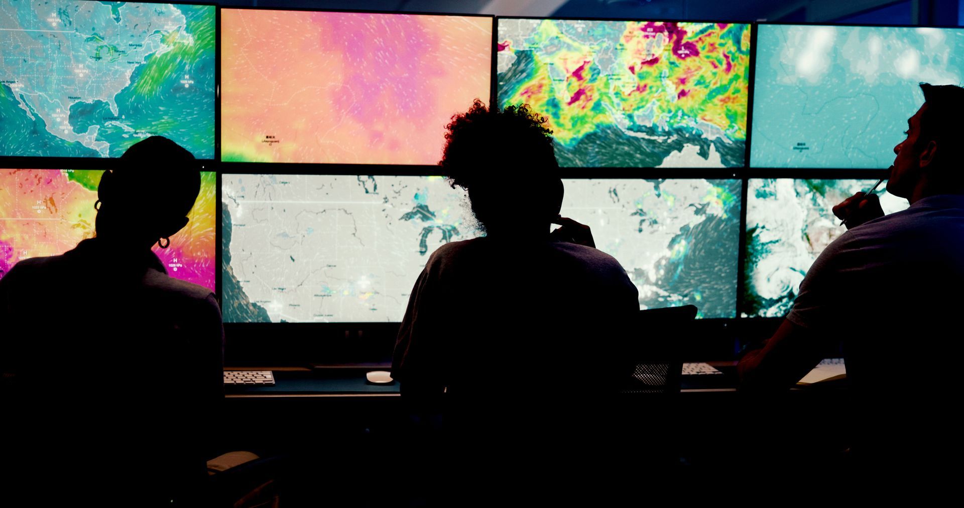 Three people monitor weather data on multiple screens in a dimly lit control room.