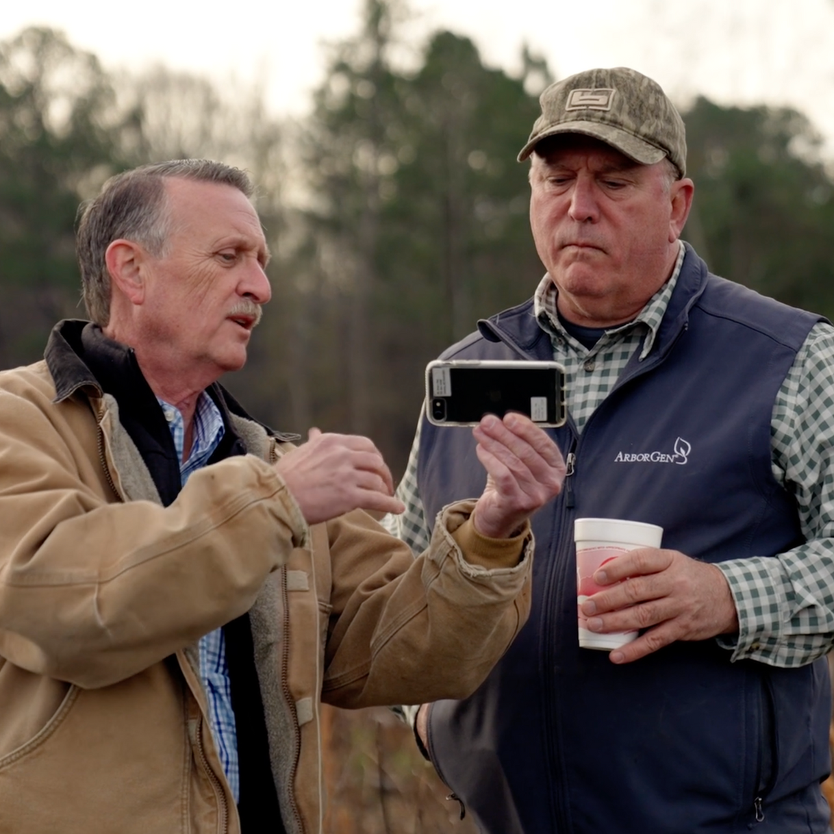 Two men looking at a cell phone with one wearing a hat that says ' georgia ' on it