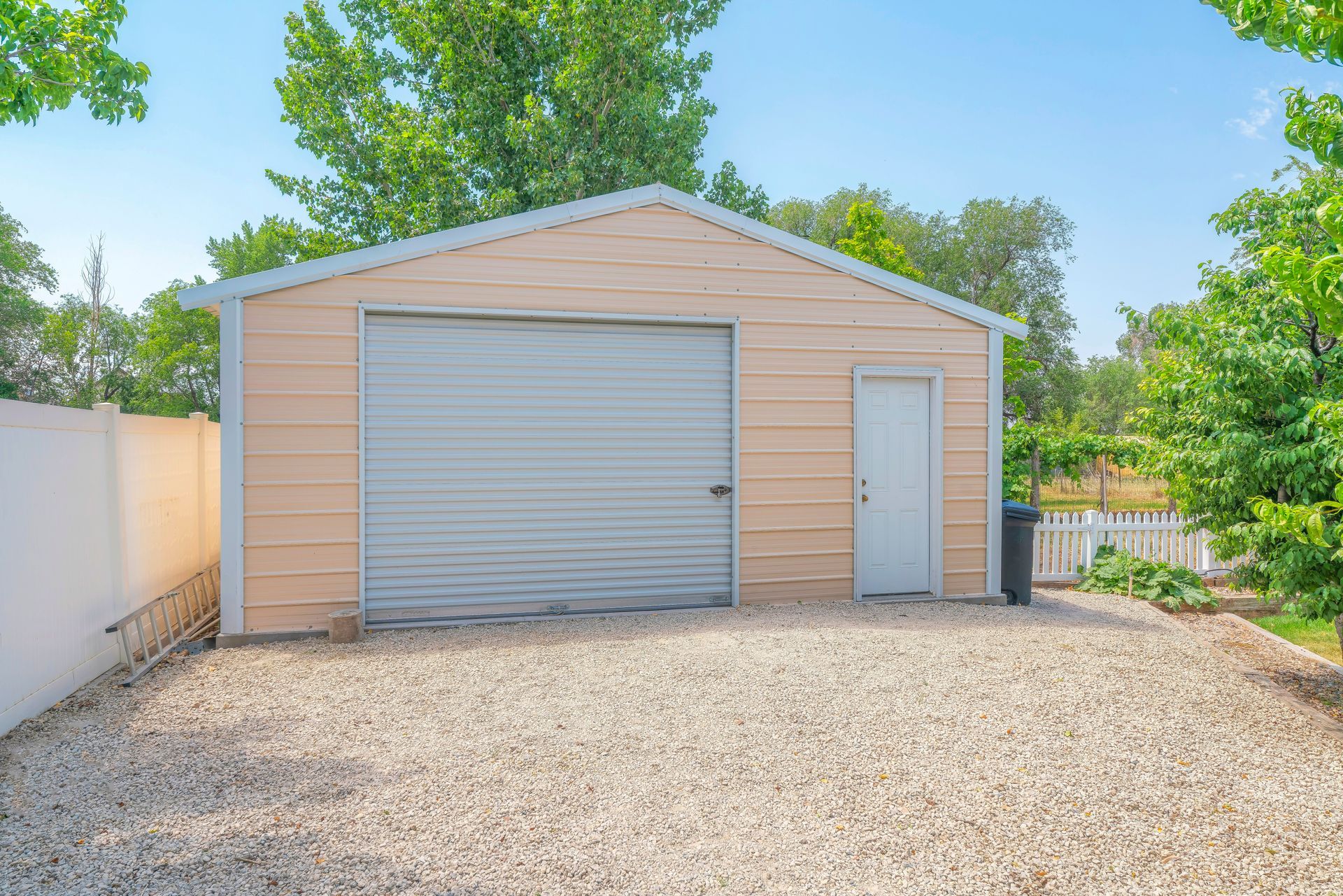 A blue garage with a white door is in the middle of a gravel driveway.