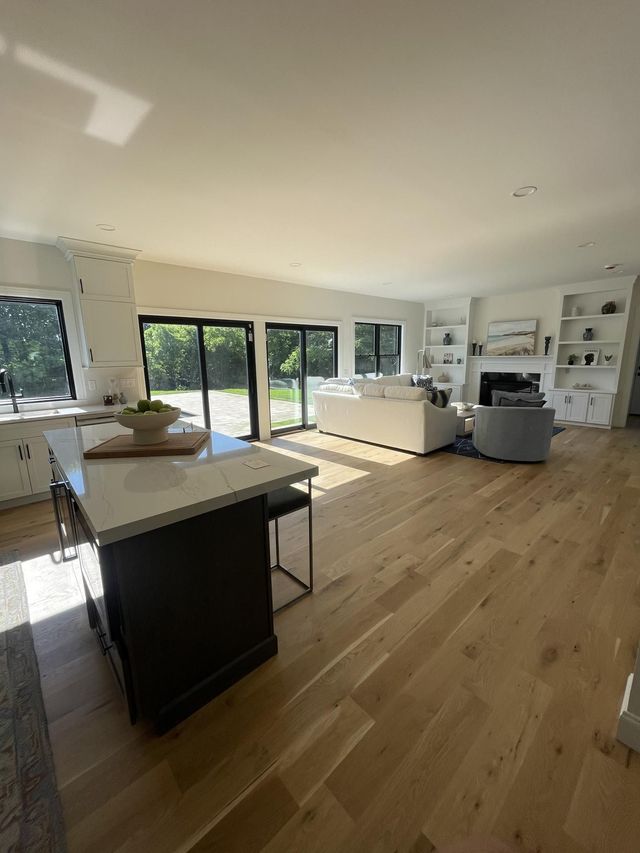 Bright, open-plan living room with wood floors, large windows, built-in shelves, and a kitchen island in the foreground