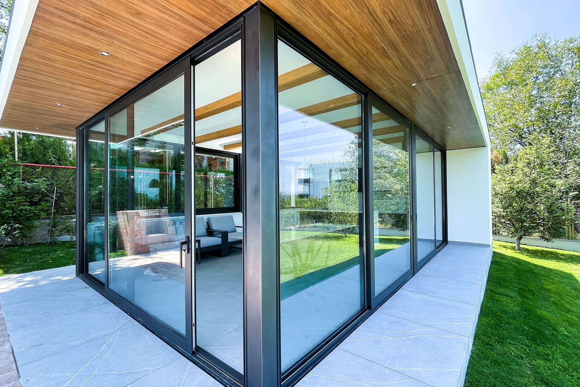 Modern glass-walled building with wood ceiling and black frames, overlooking a green yard.