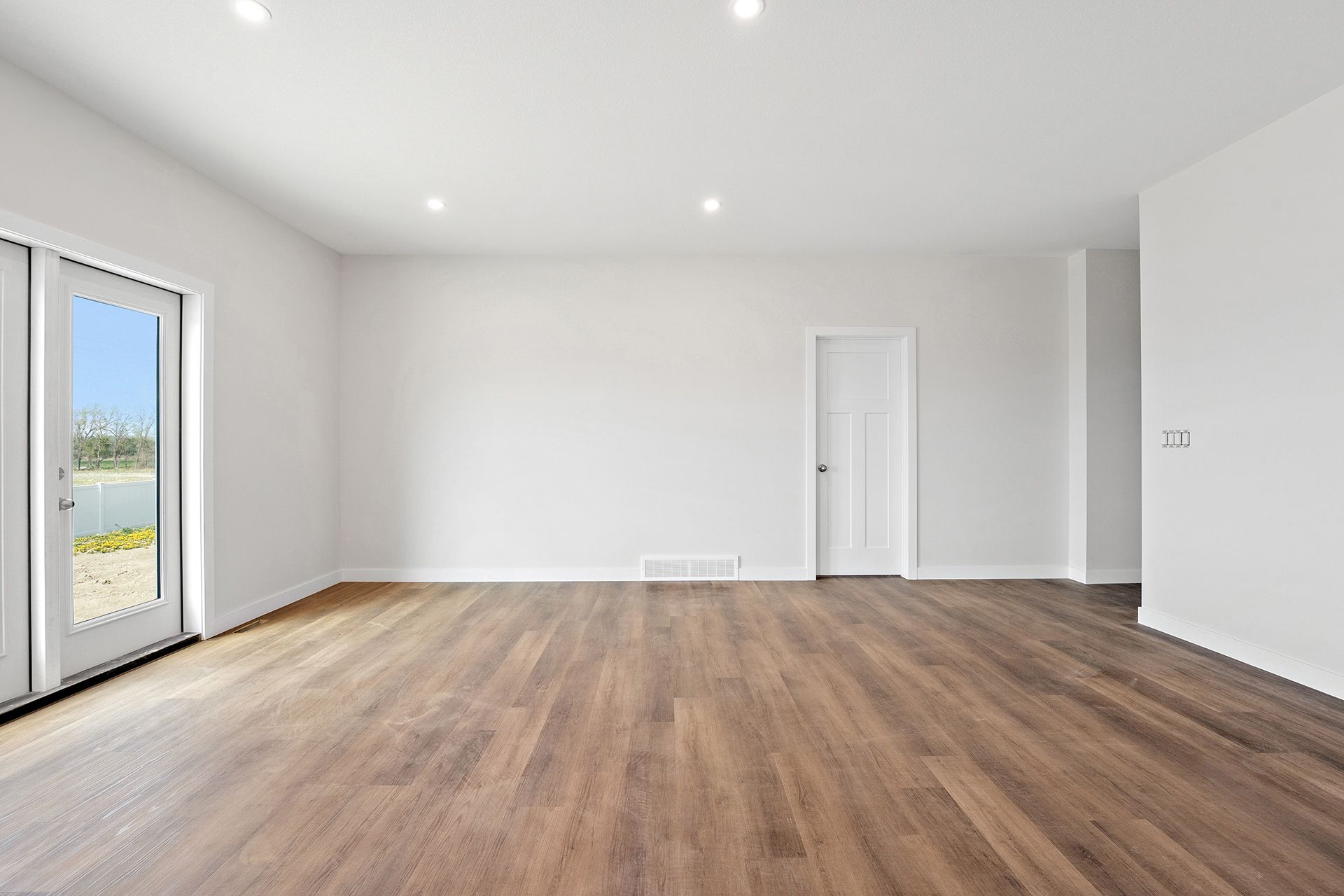 Empty room with white walls, brown wooden floor, and a door to a bright outdoor view.
