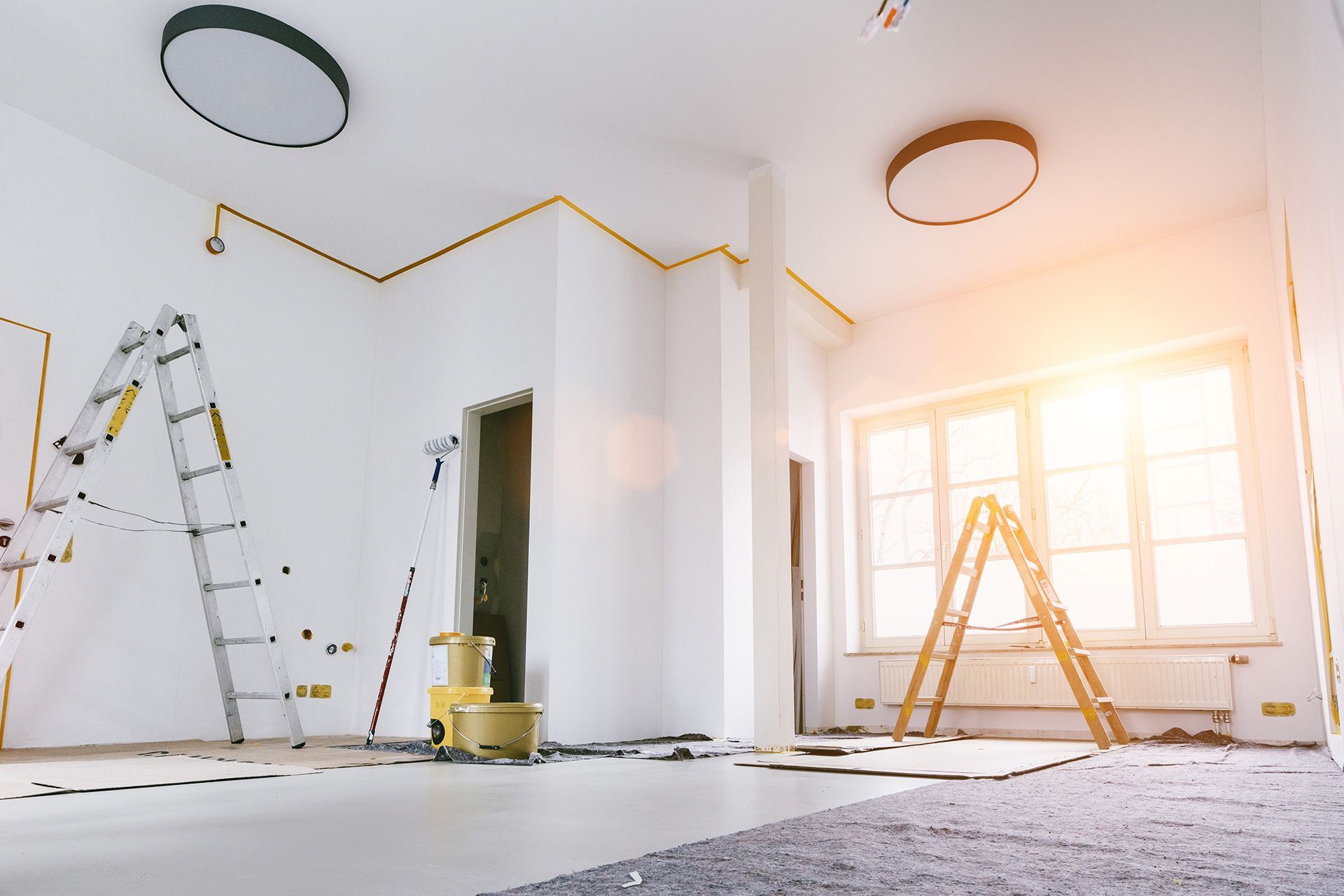 Interior of a room under renovation with ladders, paint cans, and yellow masking tape on the white walls.