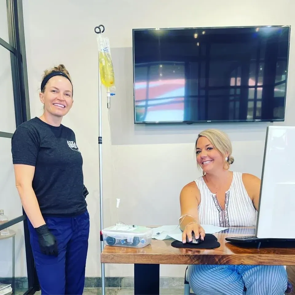 Two women in an office; one has an IV drip, the other at a desk.