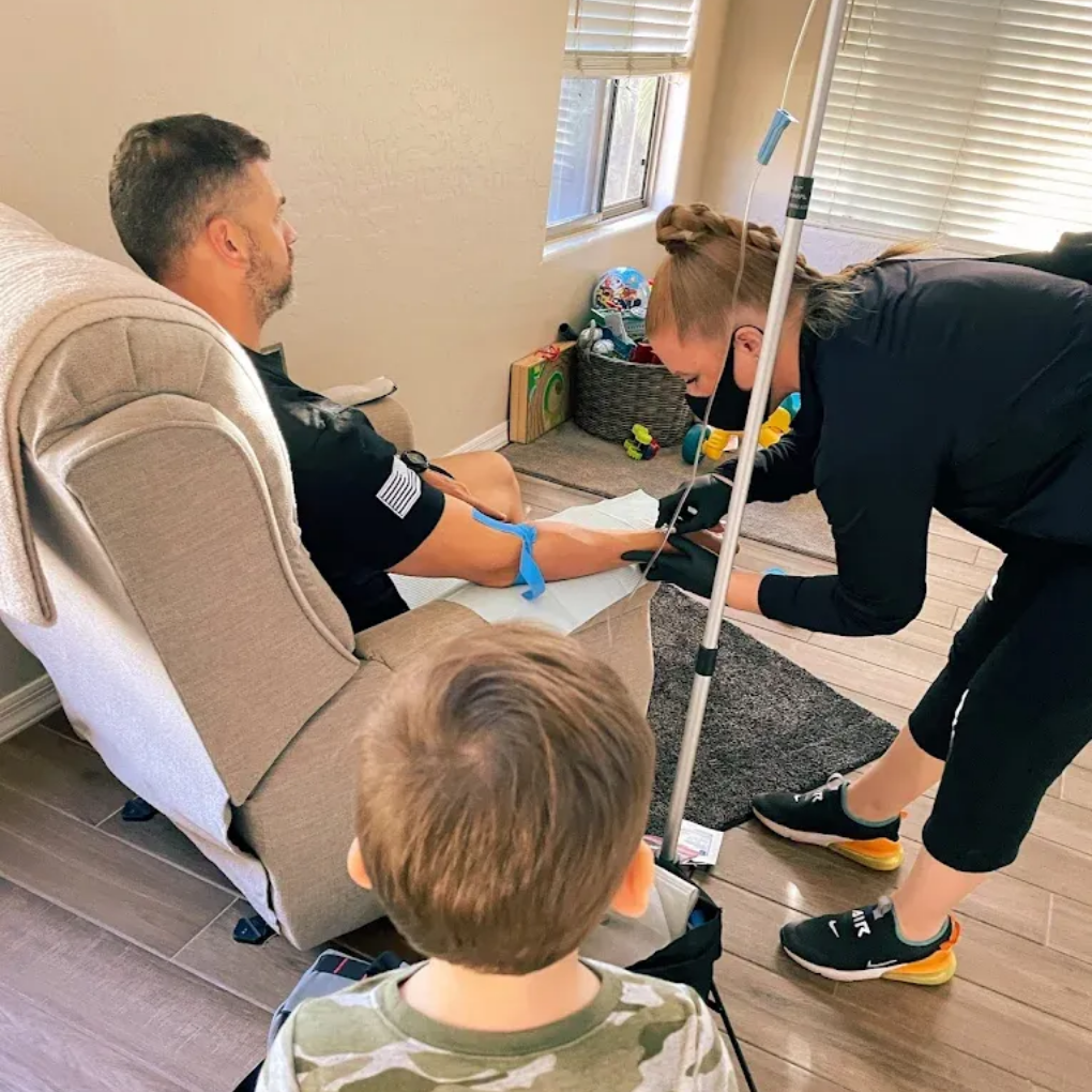 Man receiving IV therapy at home, child watches. Healthcare worker prepares equipment.