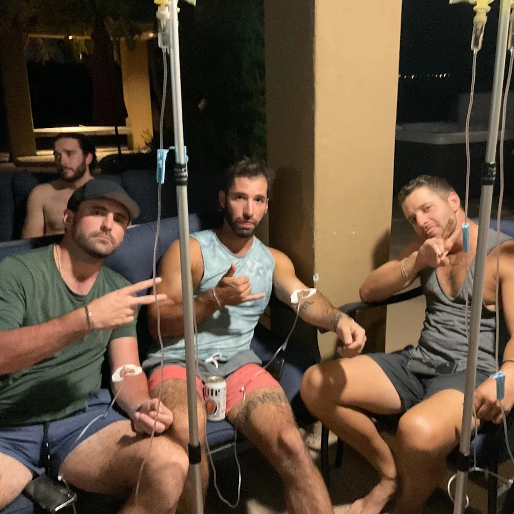 Four men on a patio receiving IV drips. One makes a peace sign, another a shaka. Miller Lite can nearby.