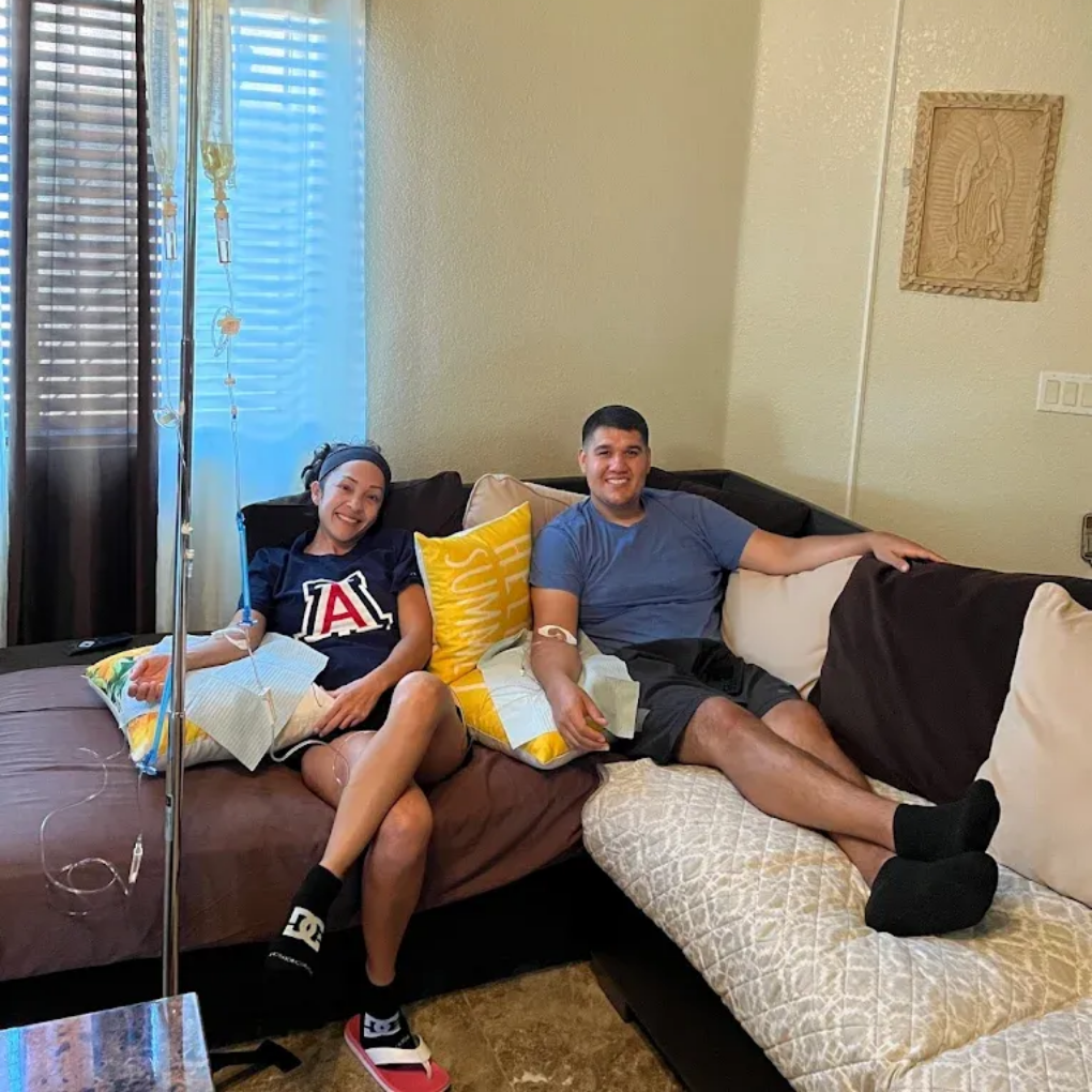 Two people on a couch with IV drips. One has a head covering. Bright room, beige and brown decor.