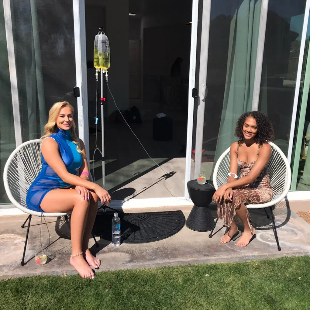 Two women sit outside, one with an IV drip. They are next to a window.