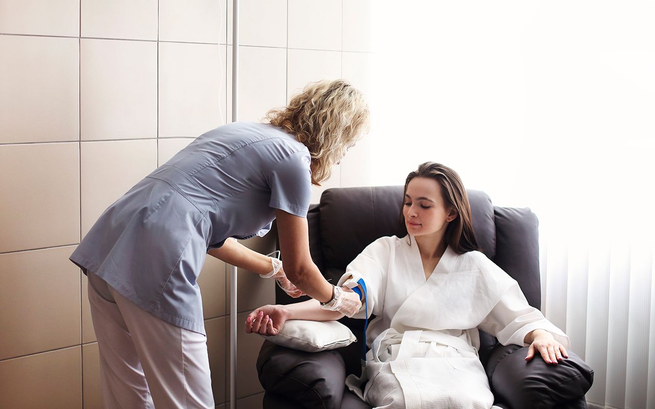 A medical professional in scrubs prepares an IV for a patient sitting in a reclining chair in a bright, modern room.